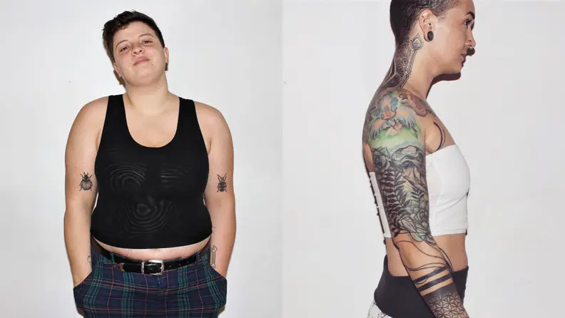 A person with their hands in their pockets wearing a black binder and jeans, next to a bald person with tattoos wearing a sleeveless white binder, facing the side.
