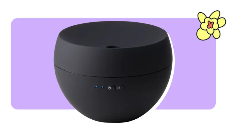 A black Stadler Form Aromatherapy Oil Diffuser in front of purple background with a yellow cartoon flower in the corner.
