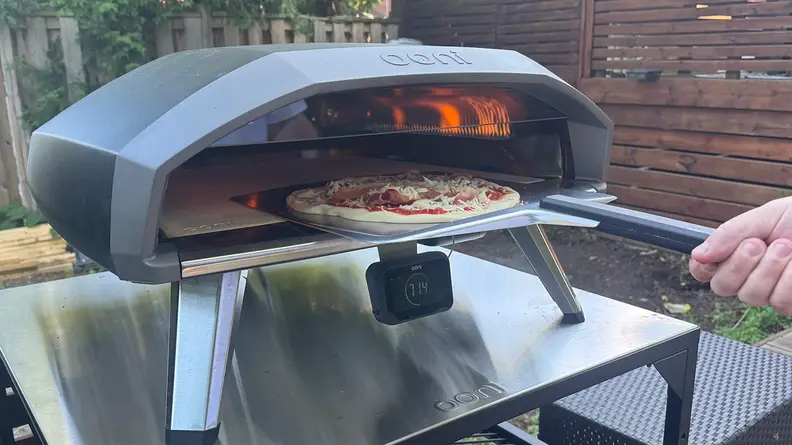 An Ooni outdoor pizza oven sits on a stainless steel table with a pepperoni pizza