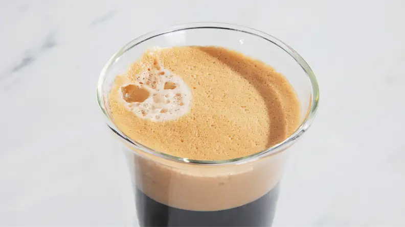 Foamy coffee in clear glass coffee mug on top of marble countertop.