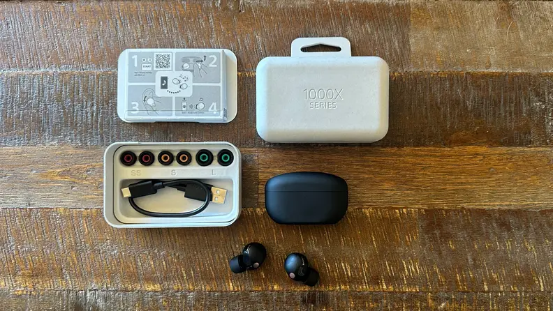 The Sony WF-1000XM5 earbuds, their case, the biodegradable packaging, and extra earbuds on a wooden table.