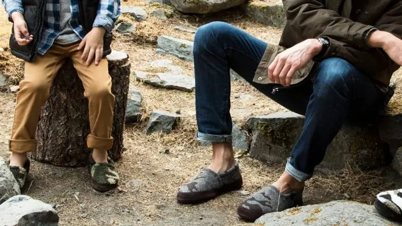 A child and a man sitting together, each wearing a pair of Acorn Moc Slippers in men's sizes.