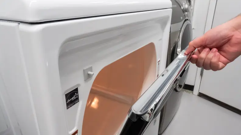 A close-up of a hand reaching into frame to open the Maytag MED7230HW dryer's hamper door, which swings from top to bottom.