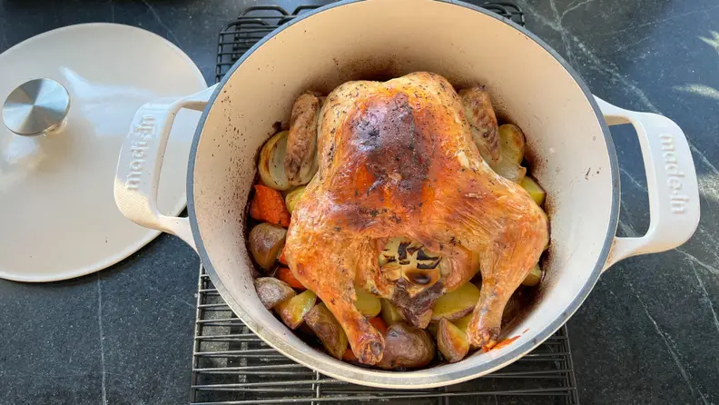 Whole cooked chicken on top of roasted veggies inside of the Made In Enameled Cast Iron Dutch Oven that is on top of a wire cooking rack.