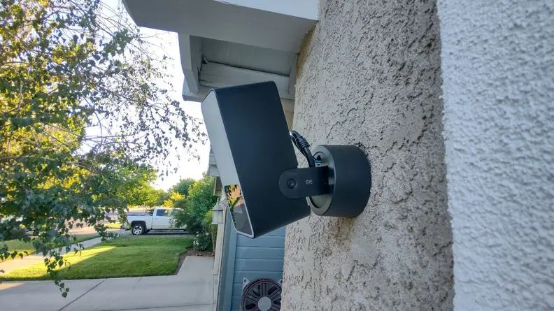 The Eve Outdoor Cam smart floodlight hands on the side of a garage wall on the exterior of a home with a neighborhood view in the background