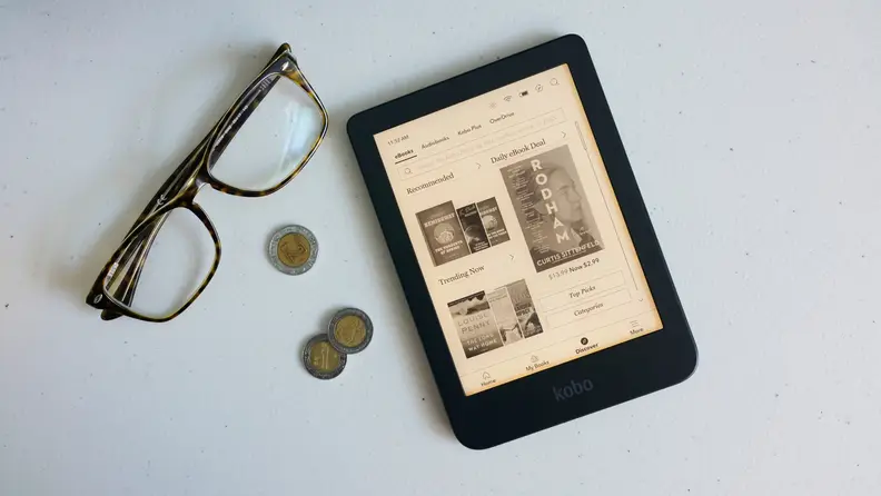 An e-reader laying flat on top of a desk next to a pair of glasses and other objects.