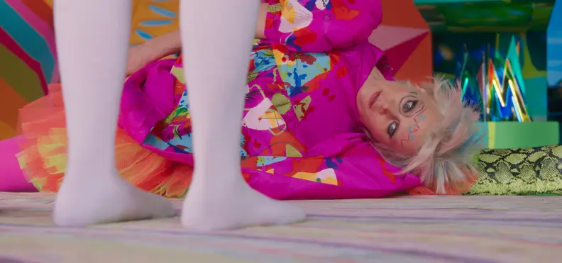 Weird Barbie, played by Kate McKinnon, lays sideways on the ground at Stereotypical Barbie (Margot Robbie's)'s feet