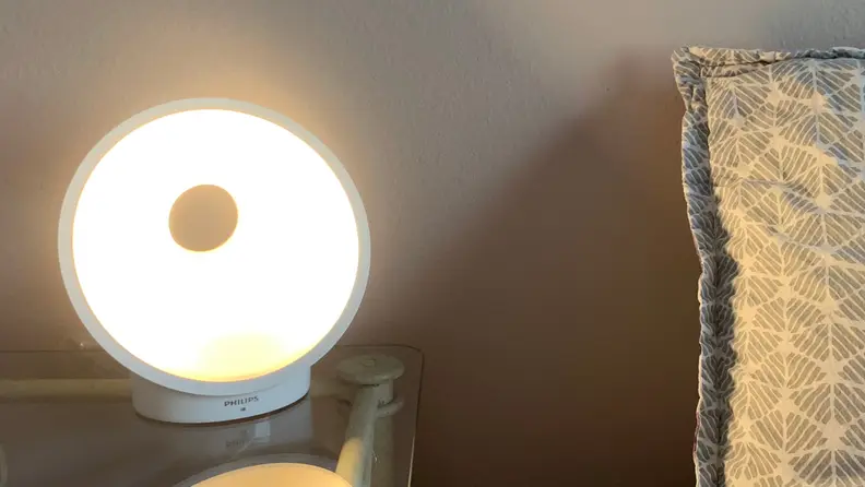 Image of the Philips Connected sunrise alarm at full brightness in a room on a clear end table next a bed with a grey pillow