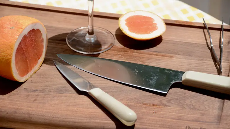 On an Our Place walnut wood cutting board, there's a half-sliced grapefruit and a wine glass. Next to these items, there's a paring knife and a chef's knife by the same brand.