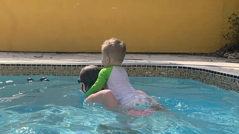 A small kid sits on a big kids back as they swim around the pool together.