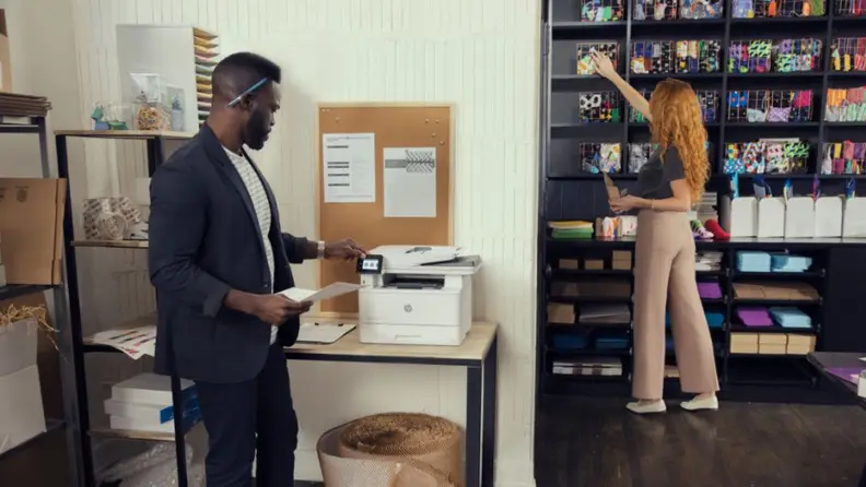 A man in a casual workspace using an HP LaserJet Pro 4101fdw while a colleague organizes a shelf in the background.
