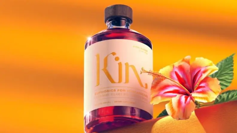 A bottle of alcohol-free elixir by the brand Kin Euphorics against a soft orange backdrop, accompanied by an orange tropical flower.
