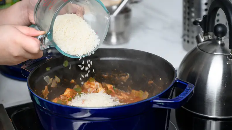 A bowl of jasmine rice being poured into a big pot of jambalaya cooking on a stove.