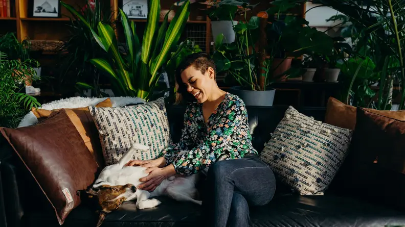 A woman pets her dog in a room of plants.