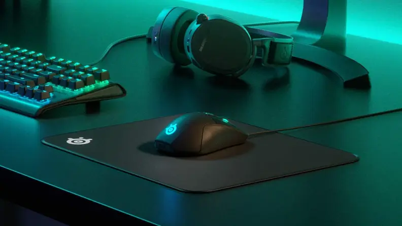 SteelSeries black mouse pad on top of desk next to gaming headset, keyboard, and monitor.