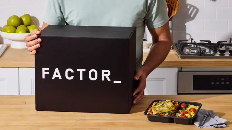 Person holding a large Factor meal delivery box in a kitchen, shown as part of a promotion using the Factor promo code.