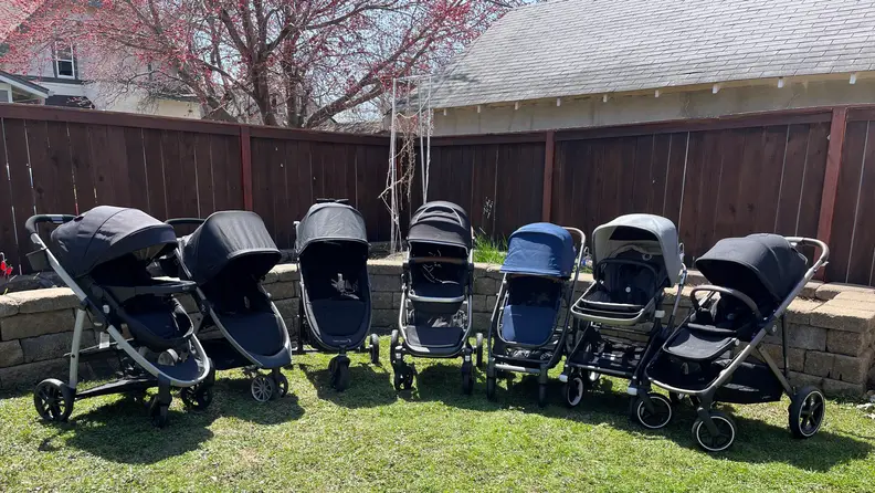 Seven different strollers lined up in a backyard.