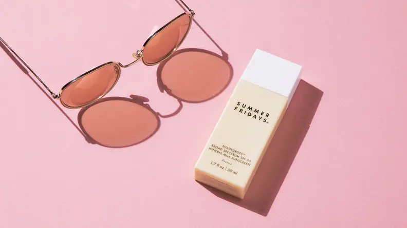 A cream-colored, rectangular bottle of sunscreen lays on a pink backdrop with sunglasses next to it.