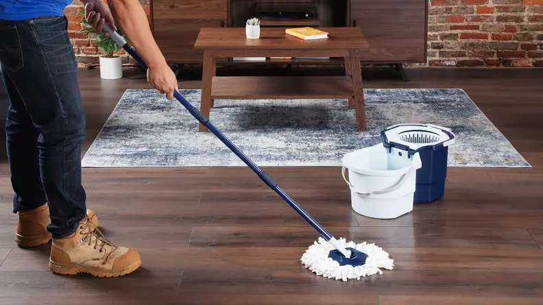 Person using Casabella Clean Water Spin Mop to clean hardwood floor surface next to bucket inside of modern living room setting.