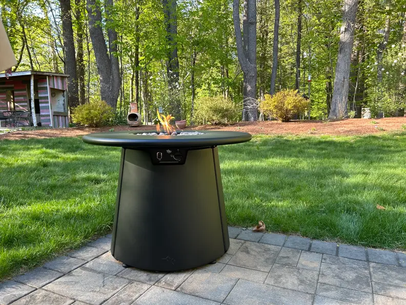 A black cylindrical fire pit sits on a patio outdoors with grass around