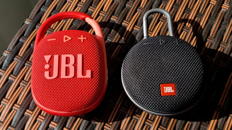 The oval shaped, brilliantly red JBL Clip 4 sits next to circular, black Clip 3 on a bar made of criss-crossed brown and black vinyl.