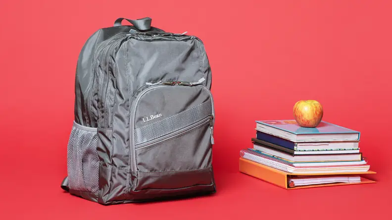 On a red background: A grey L.L. Bean Deluxe Book Pack backpack sitting next to a stack of books.