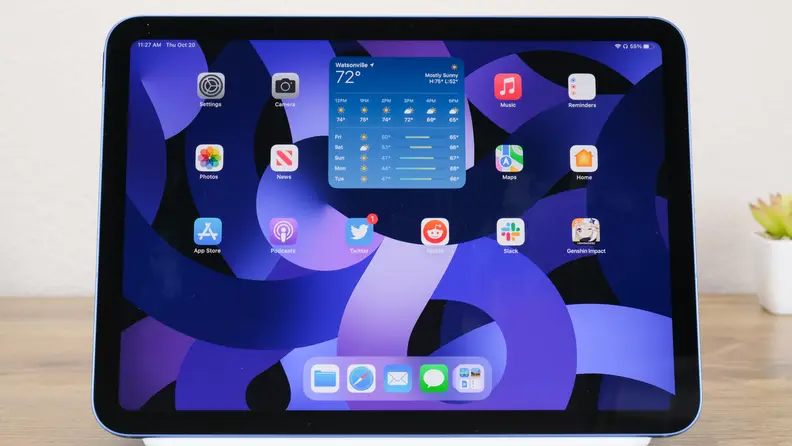 The blue Apple iPad 10 sits on a wooden desk with its home screen showing widgets.