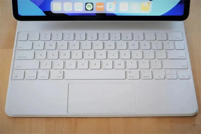 A close-up of Apple's Magic Keyboard (white), showing the layout of keys, as well as the trackpad. The bottom portion of the iPad's display is in frame, but only enough to see iPadOS's dock of apps.