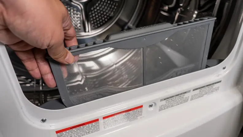 A close-up of a hand reaching into frame to remove the Frigidaire FFRE4120SW dryer's lint trap. It's about a third of the way removed in the photo.