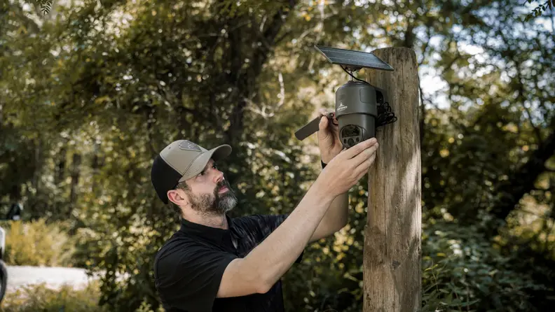 A man mounts a solar-powered camera to a wooden post in a wooded area, adjusting its angle and wiring.