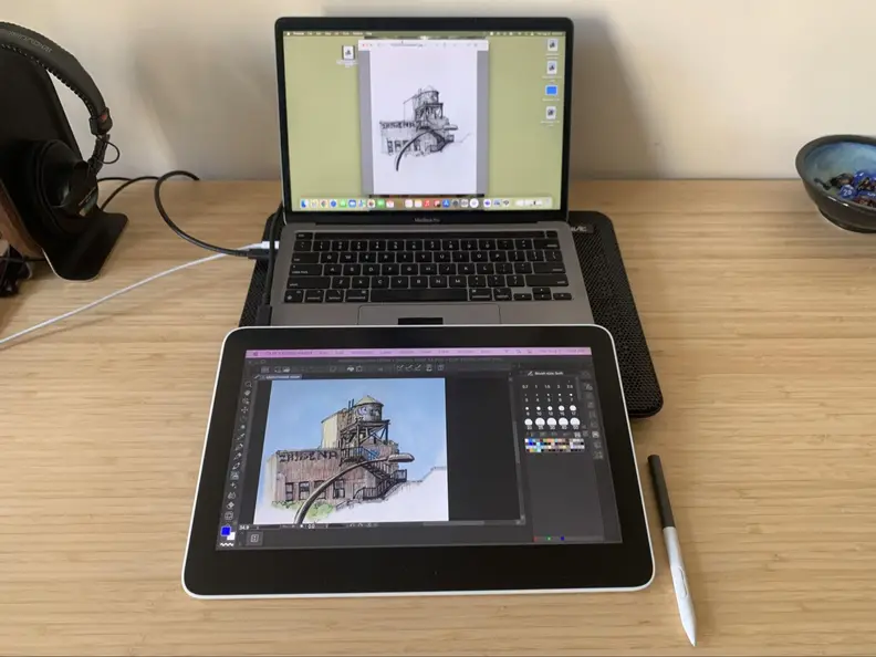 The Wacom One 13 Touch drawing tablet appears in front of a Macbook Pro on a desk with a few other desk items around it, the Wacom’s pen appears next to it.