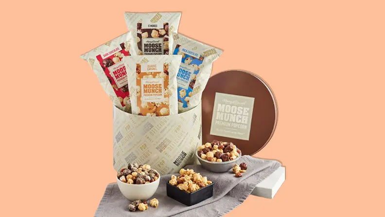 Moose Munch Premium Popcorn Classic Tin filled with popcorn packets and surrounded by three small bowls filled with popcorn on an orange background.