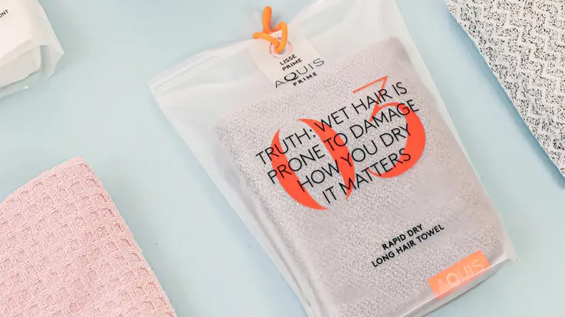 An Aquis hair towel is wrapped in its clear packaging and sits on a blue background with other towels around it. The packaging reads: "Truth: Wet hair is prone to damage how you dry it matters."
