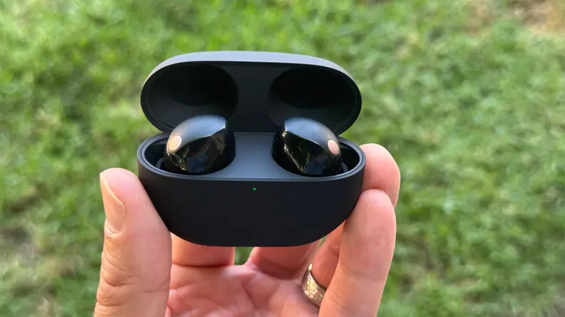The Sony WF-1000XM5 in their charing case being held in someone's fingers above green grass.