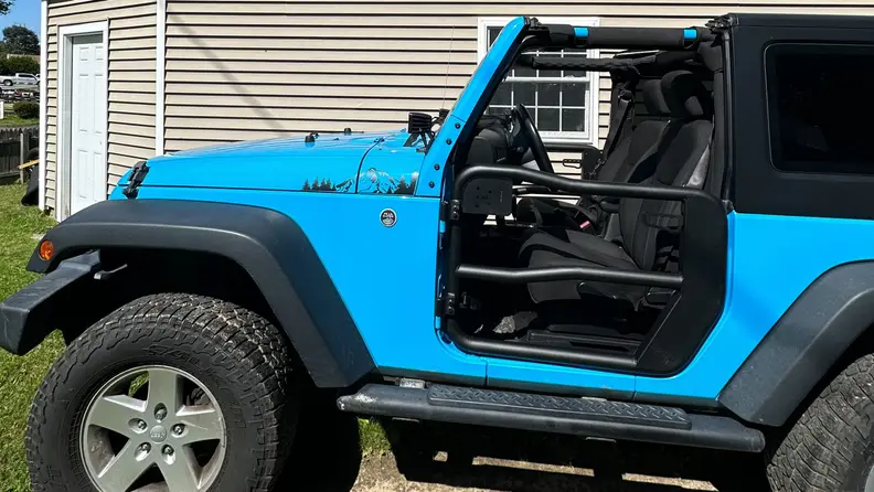 A wide profile view of a light blue Jeep with the Rugged Ridge Fortis Tube Doors installed, in front of a house.