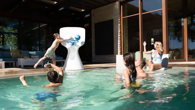 A child stands near a Ledge Lounger Hoopstr pool basketball hoop as four people watch.