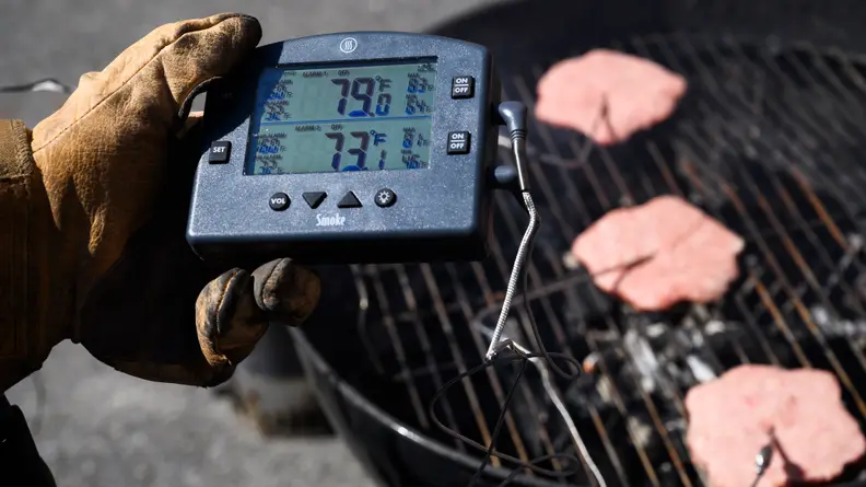 Someone holding a thermometer to check the temperature of three burgers on a grill