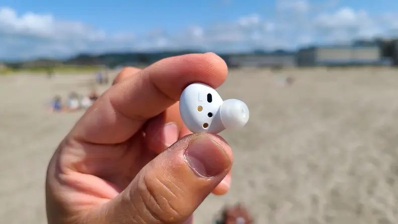 Samsung Galaxy Buds 2 is held with the interior showing against a sunny beach and mountains.