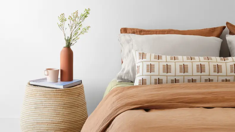 Bed with tan comforter and patterned pillows next to woven side table with a plant sitting on top