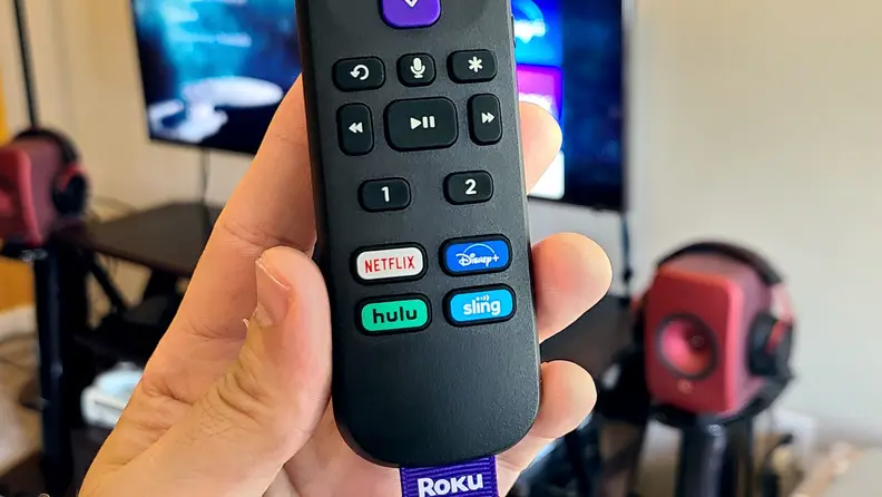 A hand holds the black, rounded remote in front of a TV console showing keys, including programmable ones as well as quick keys for streaming services.