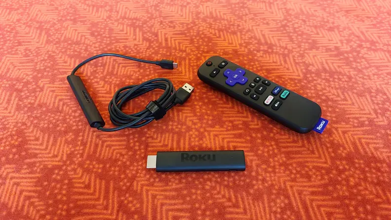The matte black, candy-bar sized Roku Streaming stick is held with its port showing above a orange-red table with accessories.