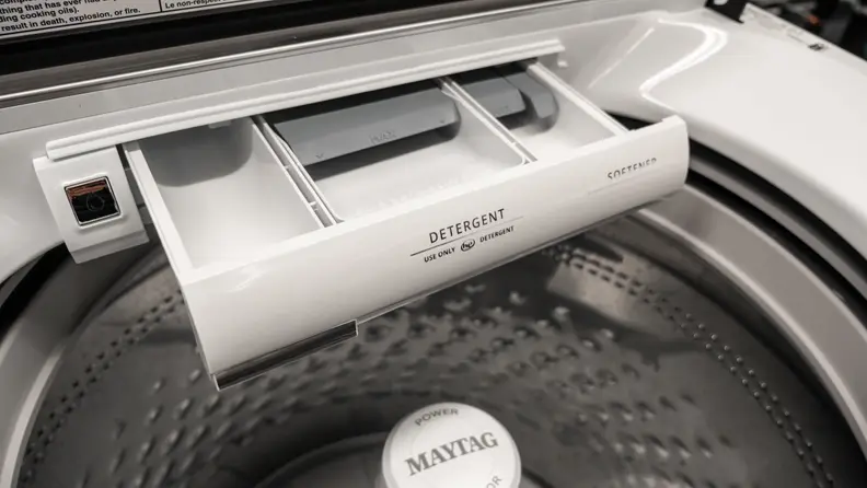 A close-up of the Maytag MVW7230HW washer's detergent dispenser, which is a small, white, plastic drawer located at the back center of the machine, under the lid but above the drum.