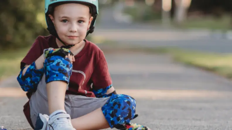 A boy sits on the ground wearing blue elbow and knee pads
