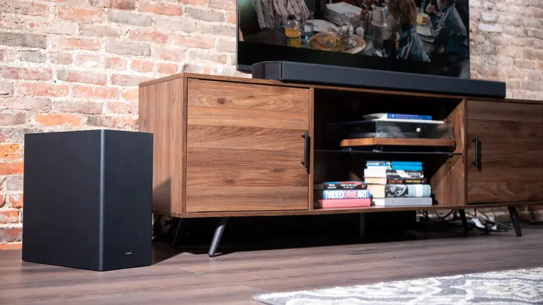 The black Q950a sits on a woodgrain cabinet in front of a large TV, with brick background.