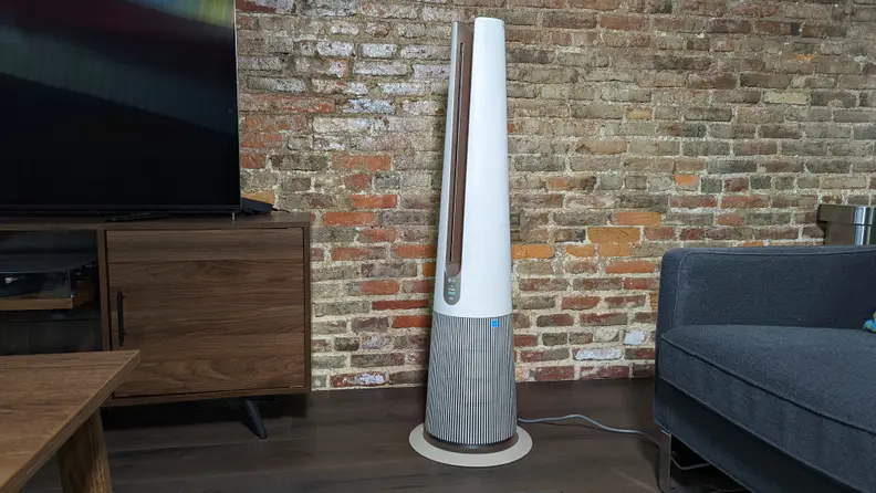 A closer view of the LG Puricare, in front of a blue armchair and next to a TV on a stand against a brick wall.