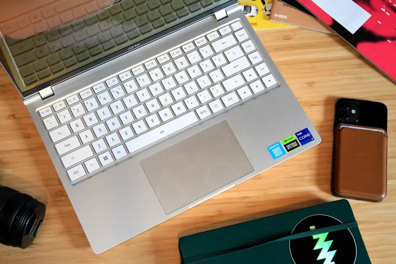 A top-down shot of a laptop with a white keyboard and silver chassis on top of a desk flanked by a few objects.