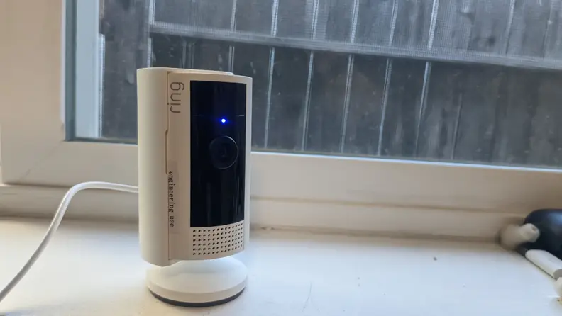 The second-gen Ring Indoor Cam with camera light on sitting on window sill in front of closed window.
