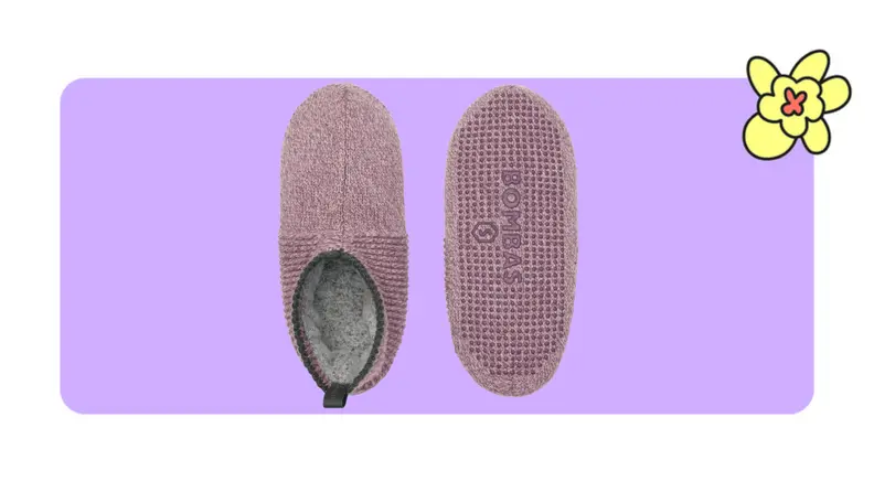A pair of Bombas slippers with fur lining in Spanish Rose color in front of a purple background with a yellow cartoon flower.