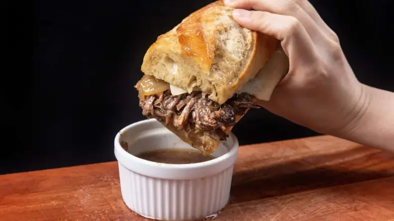 A hand holding a baguette sandwich with red meat, being dipped into a ramekin of sauce.