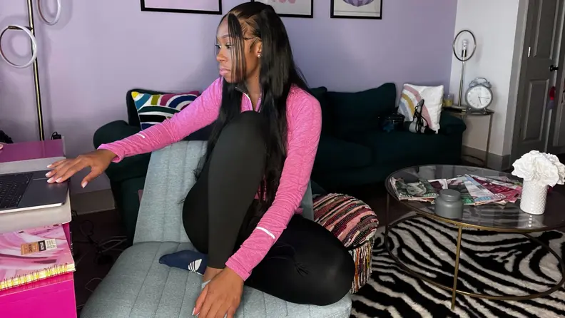 Person sitting cross legged while sitting on top of blue office chair with wide seat cushion in front of pink desk while one hand rests on laptop trackpad inside of colorful living room.
